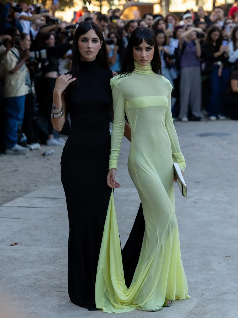 Two women in a black gown and a pale lime sheer gown outside near a crowd