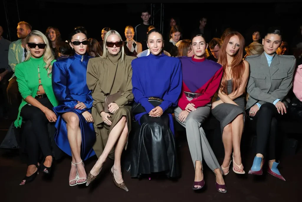 Women in bold green, blue, olive, and red outfits seated in a fashion show front row